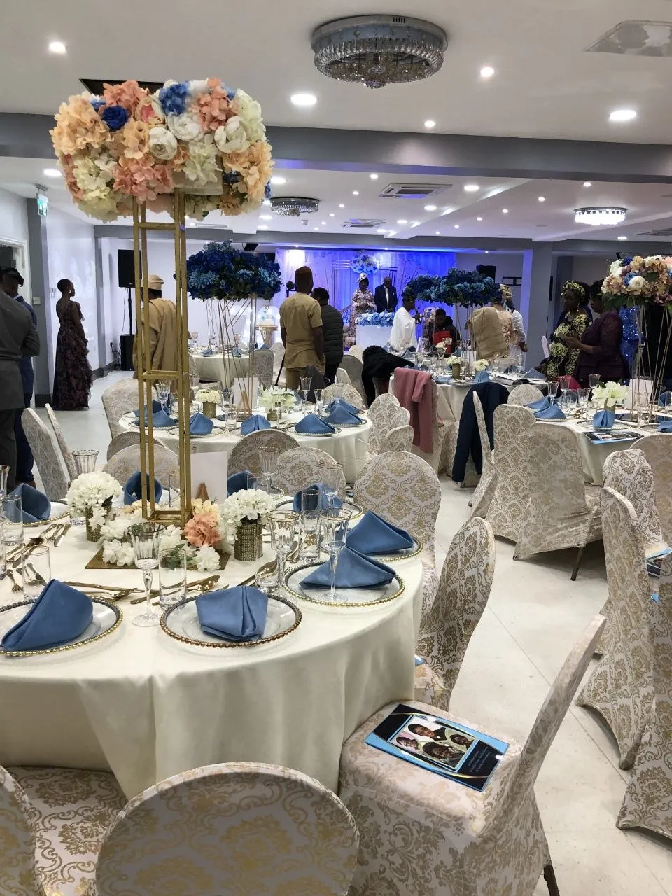 Celebration with blue napkins, gold damask chair covers and tall floral stands at P2 Centre, Erith