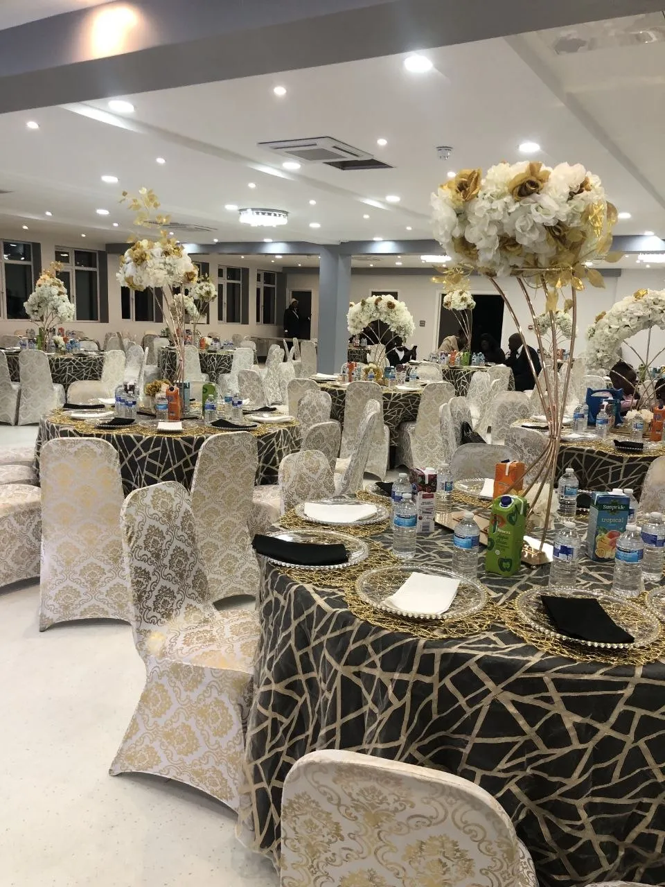 Event hall in Erith with black and gold geometric tablecloths, gold damask chair covers and tall white and gold floral centrepieces