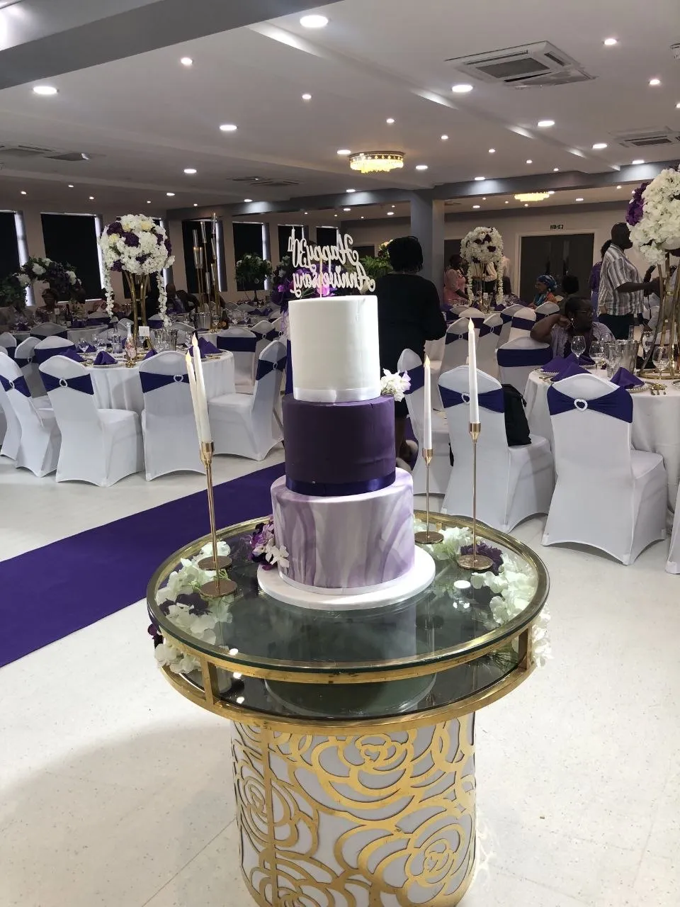 Anniversary cake in front of round tables with purple sashes in the P2 Centre hall, Erith