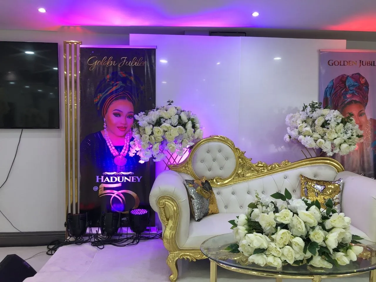 Golden jubilee 50th birthday stage with white and gold sofa, ivory roses and portrait banners at P2 Centre, Erith